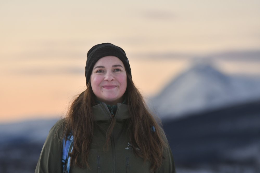 PItkähiuksinen nainen hymyilee musta pipo päässään ja katsoo kameraan. Naisella on pipo päässään ja reppu selässään, taustalla on vuori ja auringonlaskun värjäämä taivas