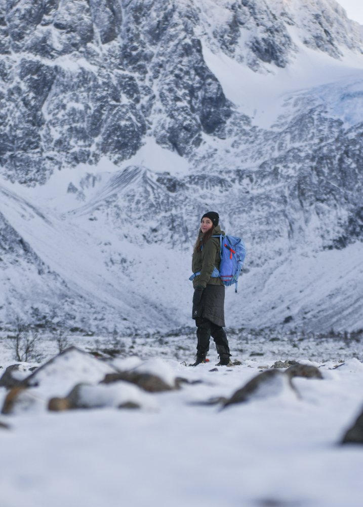 Pitkähiuksinen nainen seisoo talvivaatteissa lumisen vuorimaiseman edessä