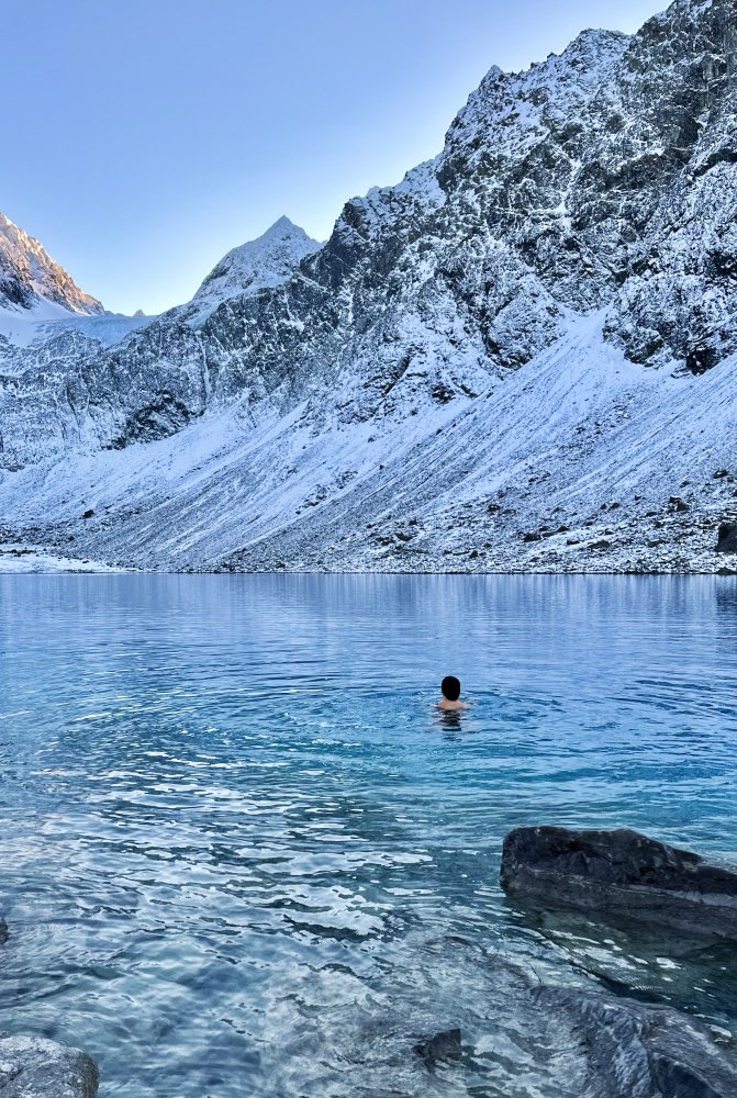 Nainen ui turkoosivetisessä jäätikkölammessa lumenpeittämien vuorten keskellä