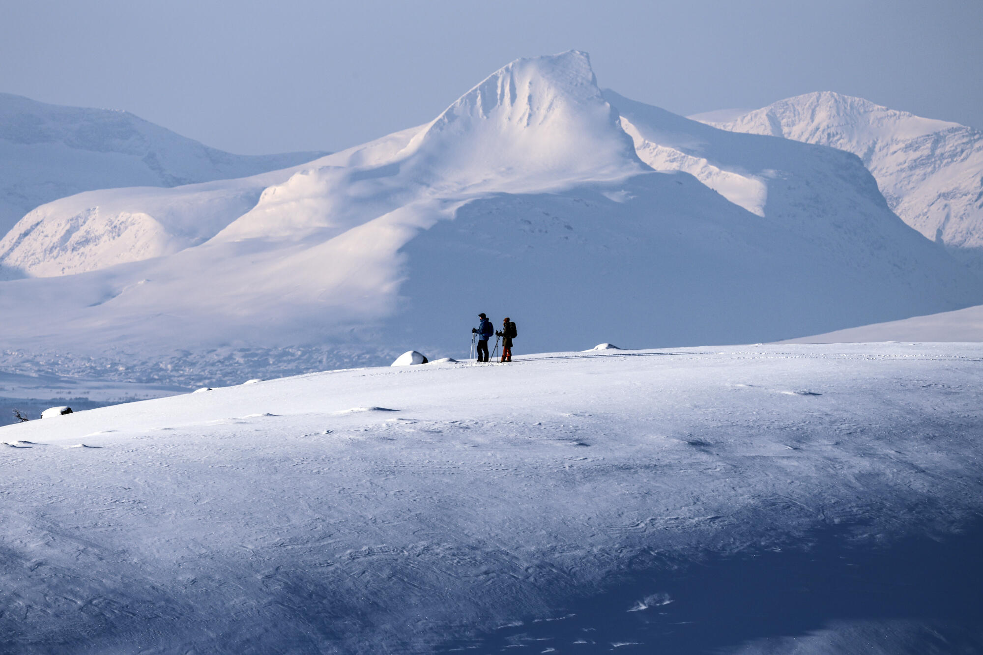 Kilpisjärven vuoristomaisemassa pariskunta hiihtää kaukaisuudessa aurinkoisena kevätpäivänä. Taustalla on Norjan Barras tunturi.