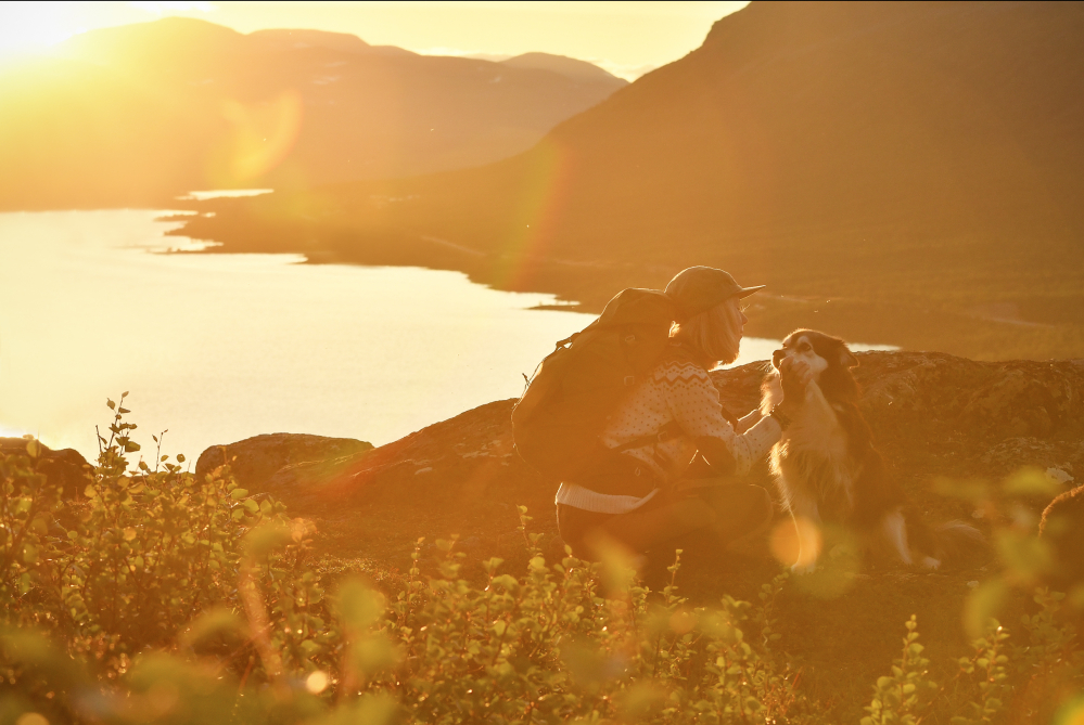 Nainen ja koira tunturin huipulla laskevan auringon valossa