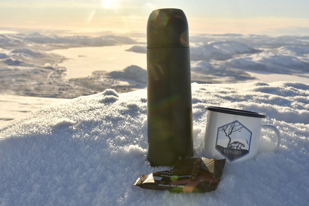 Termospullo ja Kilpisjärvi-aiheinen emalimuki lumisen kiven päällä Saanatunturin huipulla