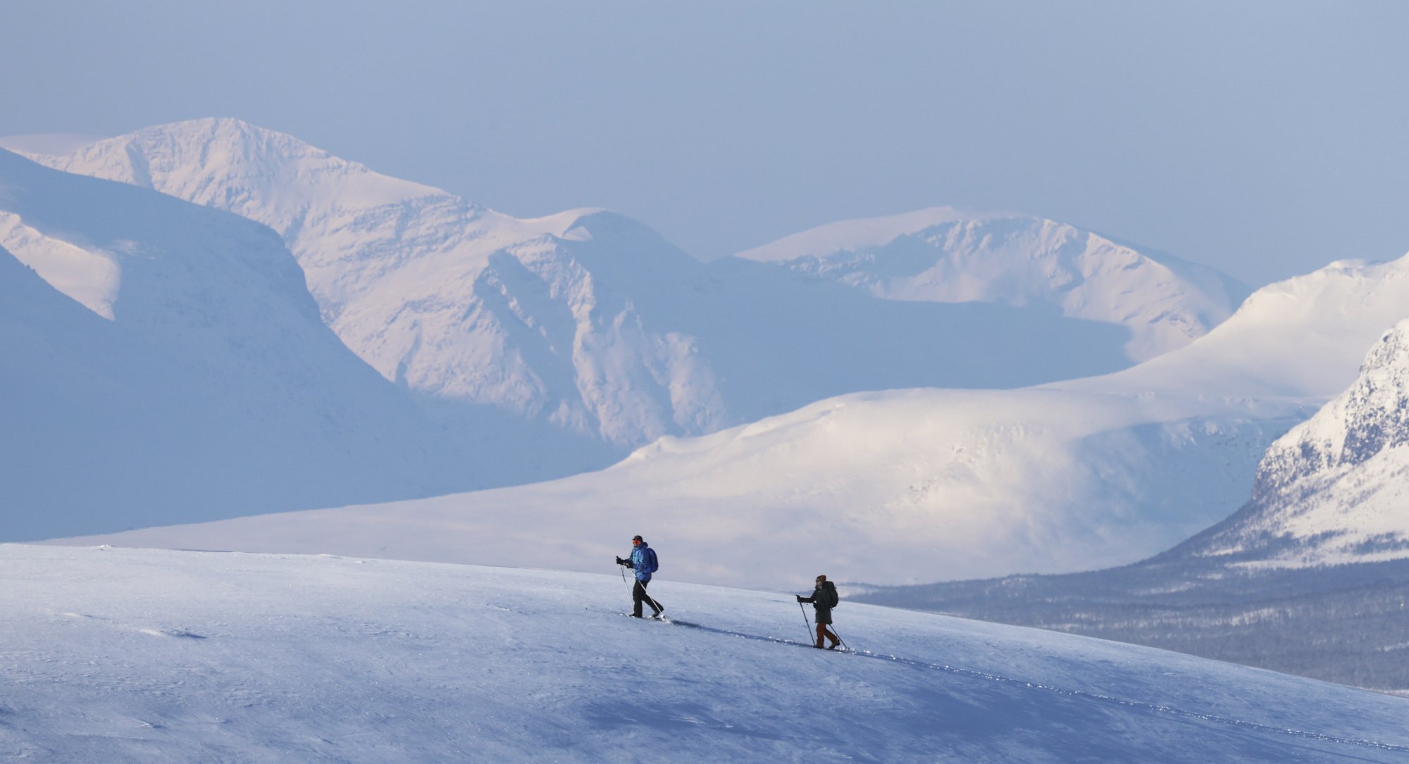 Jylhä talvinen maisema maisema Kilpisjärvellä vuorineen. Keskellä kuvaa on kaksi hiihtäjää Kilpisjärven Retkeilykeskuksen vuokravälineillä hiihtämässä mäkeä ylös