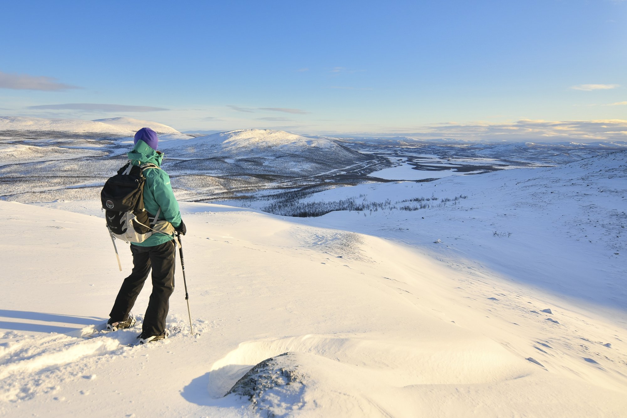Nainen katselee Muotkatakan Leutsuvaaran huipulta etelään talvista tunturimaisemaa. Hänellä on Kilpisjärven Retkeilykeskuksen välinevuokraamosta vuokratut lumikengät ja sauvat