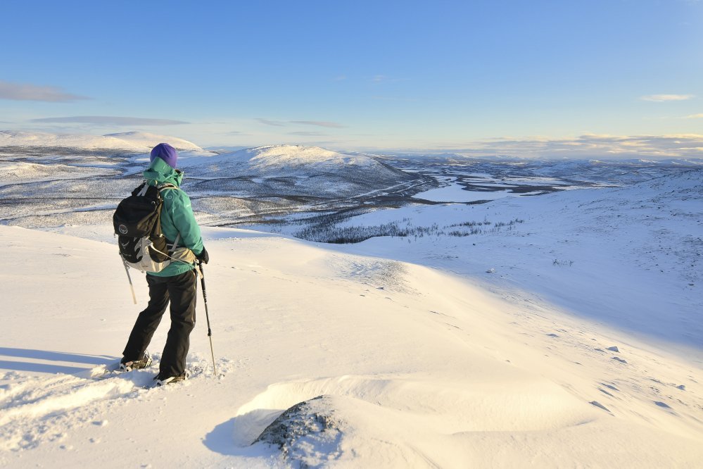 Nainen seisoo lumikengillä ja pitää sauvoista kiinni samalla kun katselee talvista maisemaa Leutsuvaaran laidalta etelään kohti lumista laaksoa