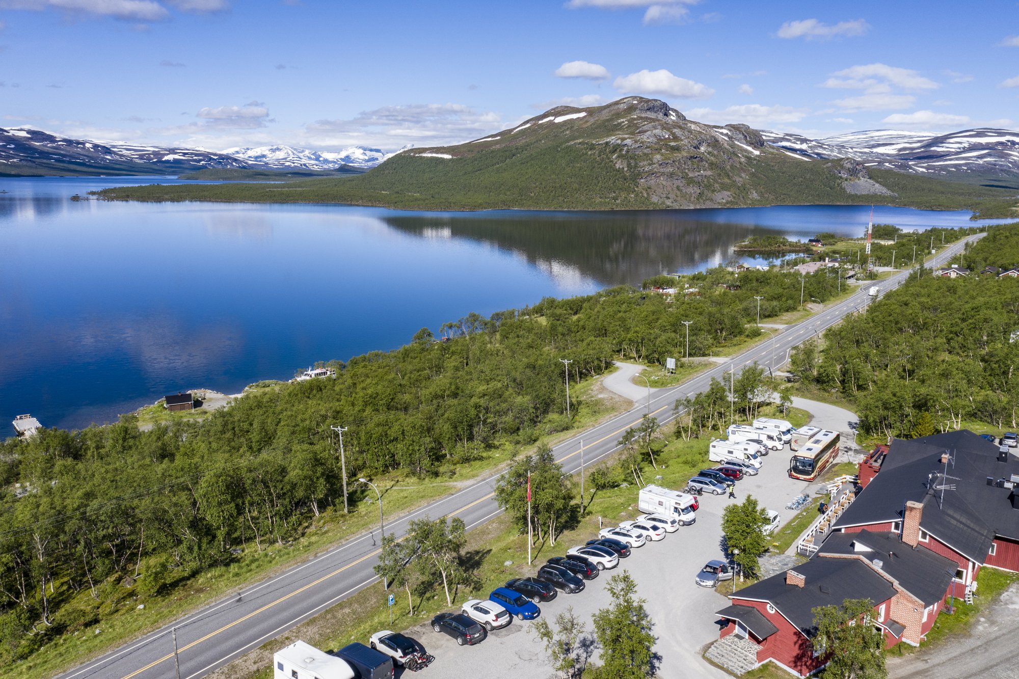 Ylhäältä päin kuvattu Kilpisjärven Retkeilykeskus kesällä, parkkialue täynnä autoja. Taustalla Kilpisjärvi ja vielä vähän luminen tunturimaisema.