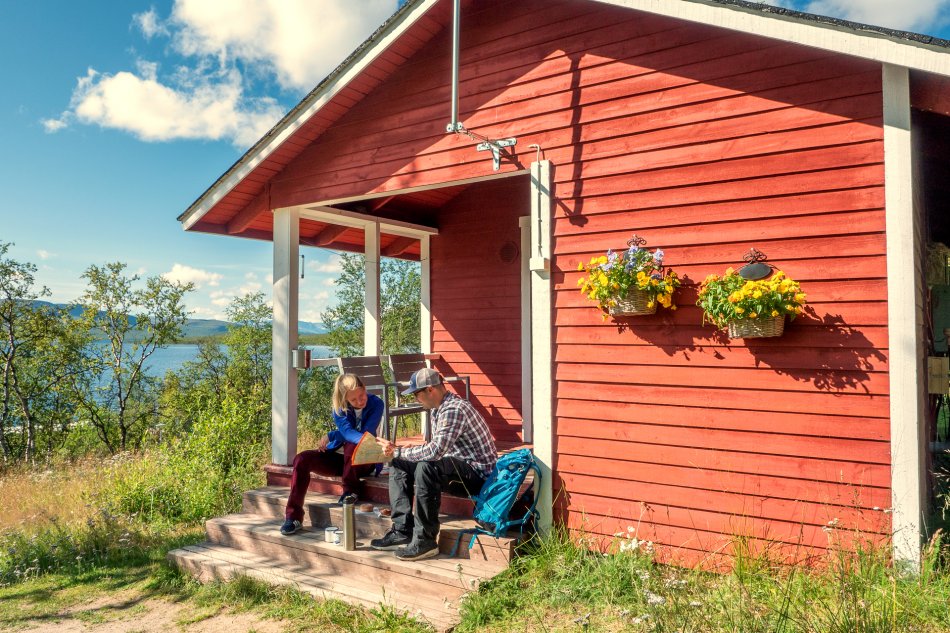 Punainen mökki Kilpisjärven rannalla kesäisessä auringonpaisteessa. Mies ja nainen istuvat leirintämökin portailla tutkien karttaa. Mökin seinällä on orvokkeja kahdessa ruukussa.