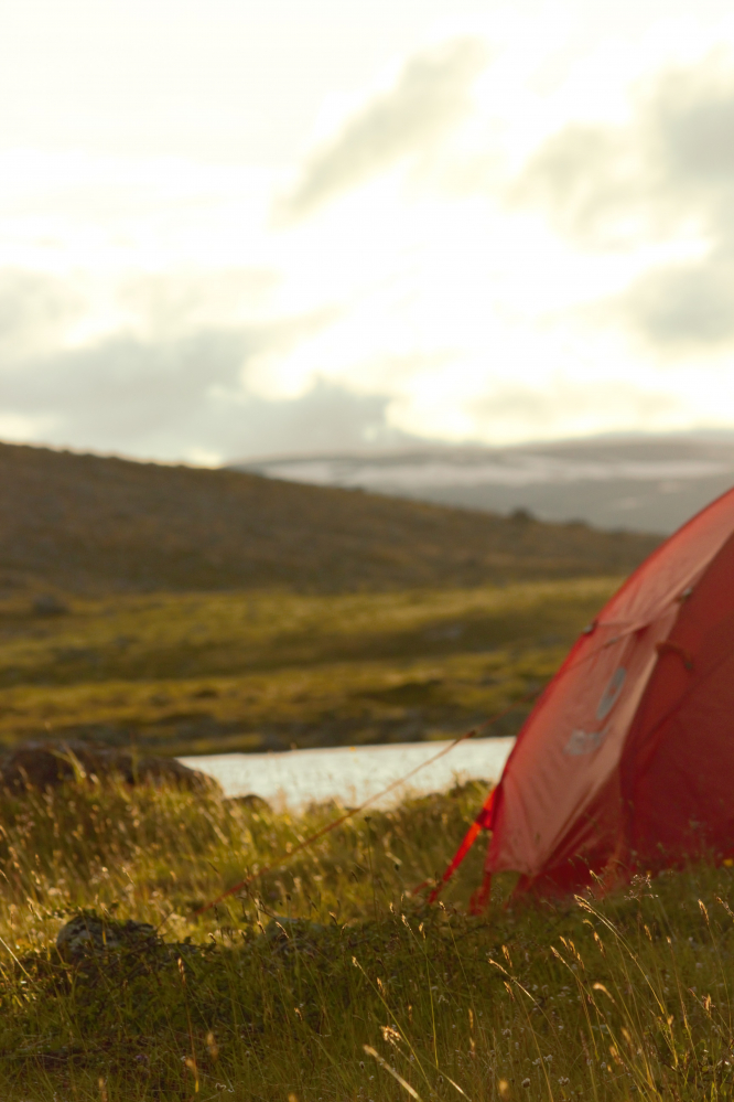 Kuvassa näkyy osa punaista telttaa tunturin rinteellä laskevan auringon laskiessa kesäiltana