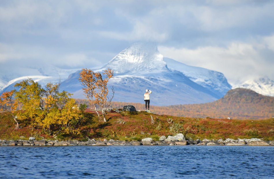 Kuvassa on värikäs maisema Kilpisjärven ruskassa, etualalla on Kilpisjärven aaltoja, keskellä ruskainen niemi, jolla seisova nainen katsoo edessään olevaa lumihuippuista Barras tuntusa