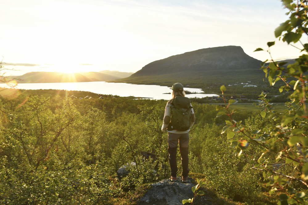 Nainen katsoo kohti auringonlaskua seisoen kivellä vaellusvaatteissa ja reppu selässään. Taustalla on Saanatunturi sekä Kilpisjärven kylä.