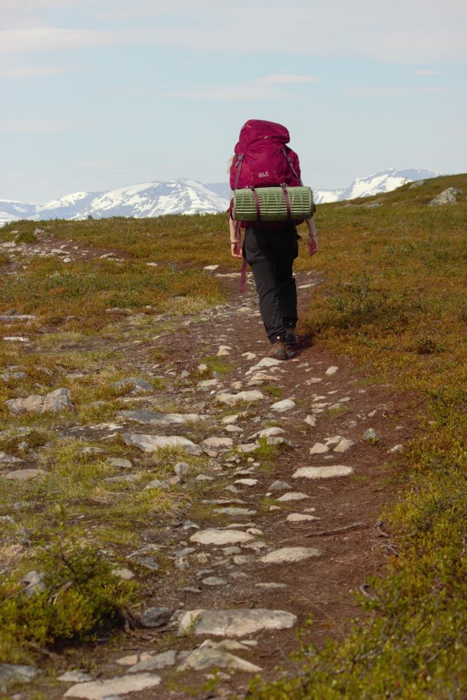 Ihminen kävelee selin kameraan kivikkoista polkua punainen rinkka selässään. Taustalla näkyy Norjan lumihuippuisia vuoria. 