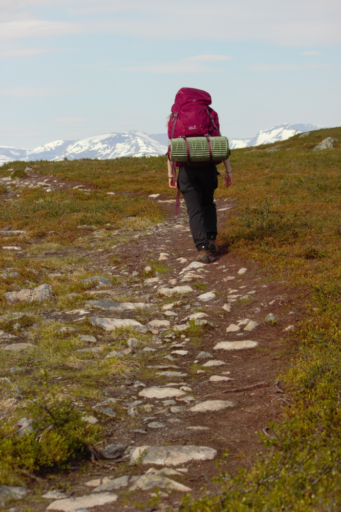 Ihminen kävelee selin kameraan kivikkoista polkua punainen rinkka selässään. Taustalla näkyy Norjan lumihuippuisia vuoria. 