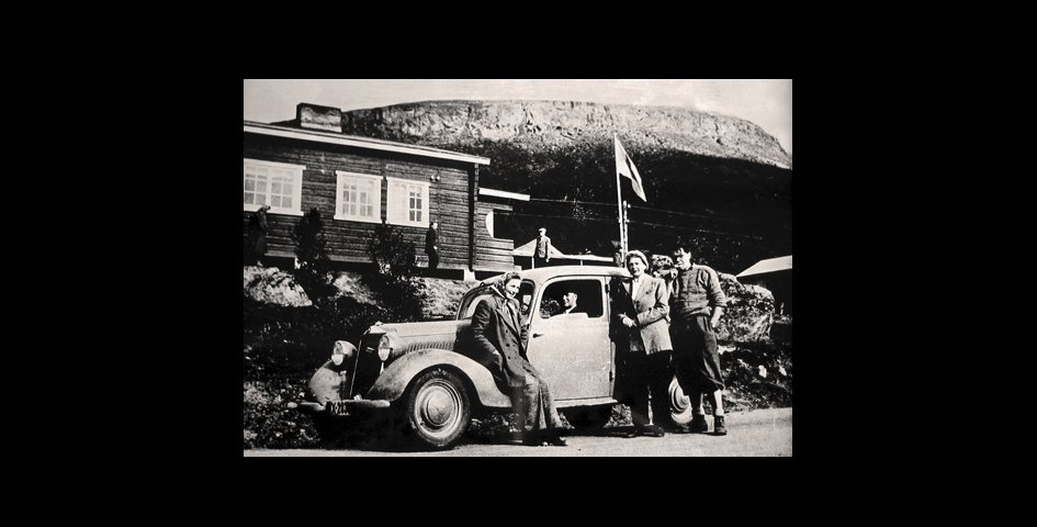 Vanha mustavalkoinen kuva Kilpisjärven Retkeilykeskuksen pihasta. Vanhaan autoon nojailee kolme ihmistä ja Saana on taustalla