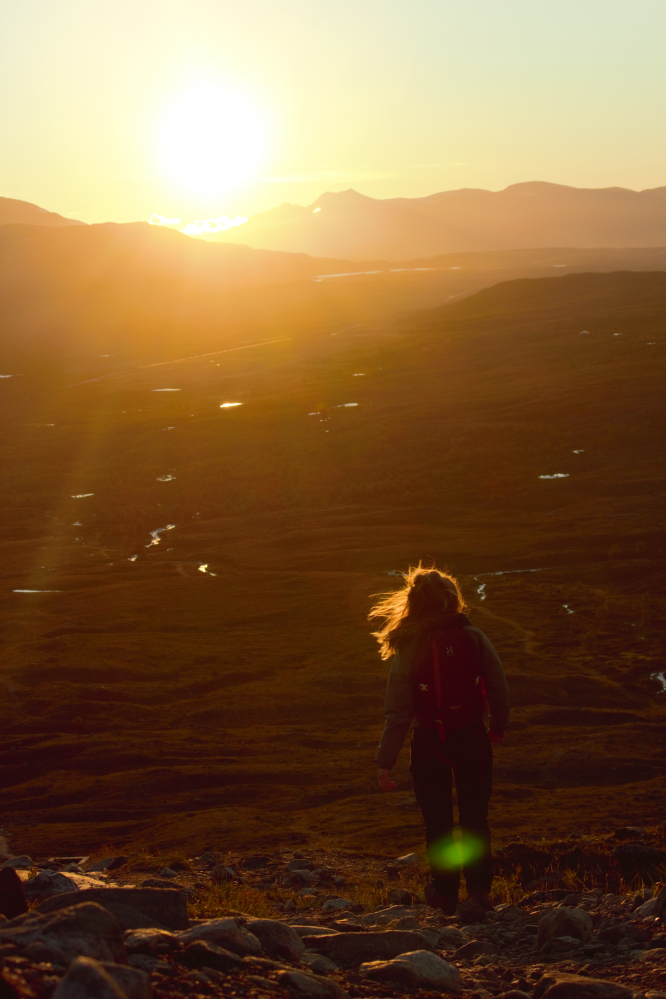 Nainen kävelee alas tunturin rinnettä kohti auringonlaskua ja taustalla näkyviä Norjan tuntureita