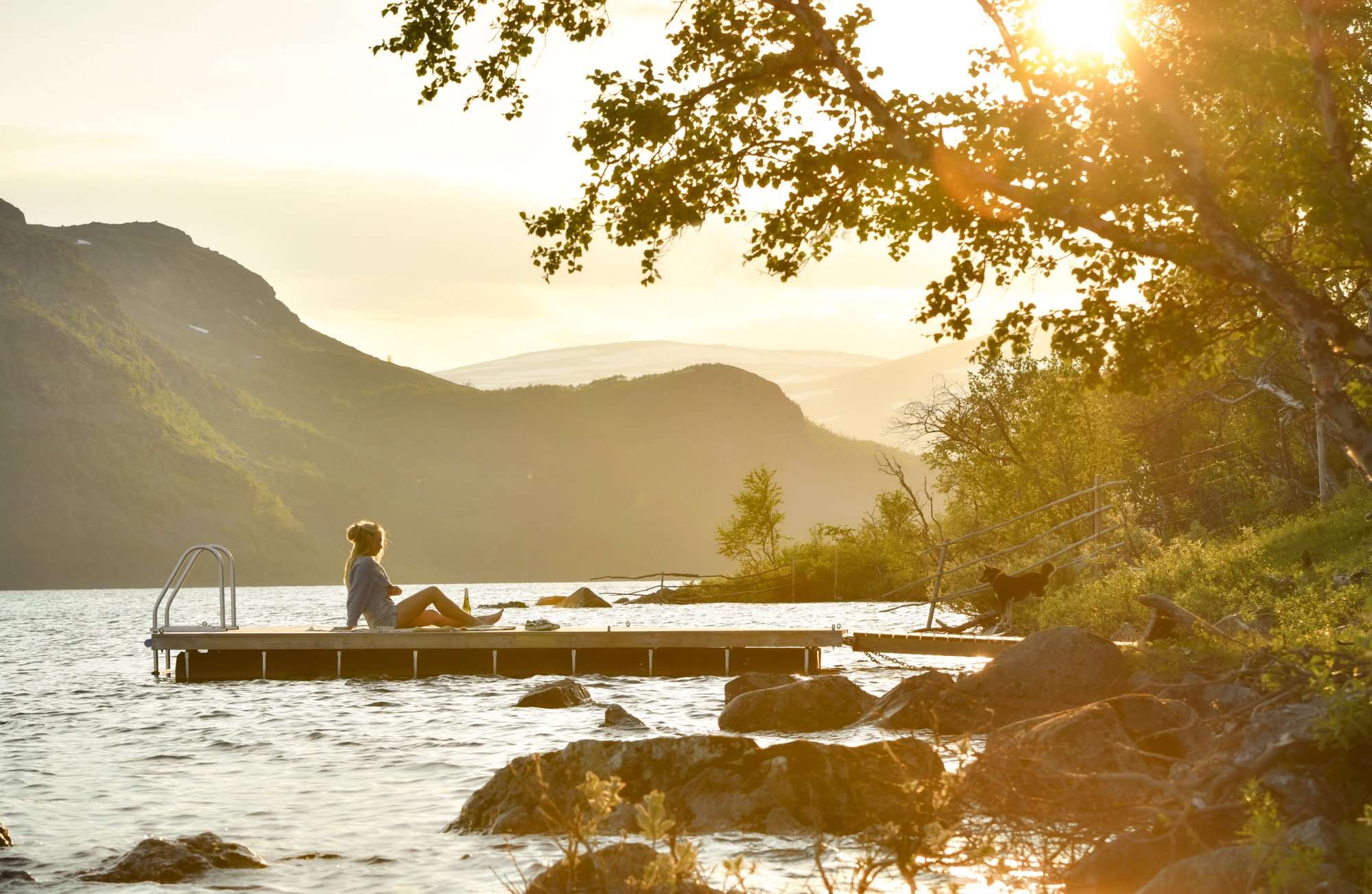 Lempeä keskiyön aurinko valaisee Kilpisjärven retkeilykeskuksen rannassa olevaa laituria, jolla nainen nauttii kesäillan lämmöstä