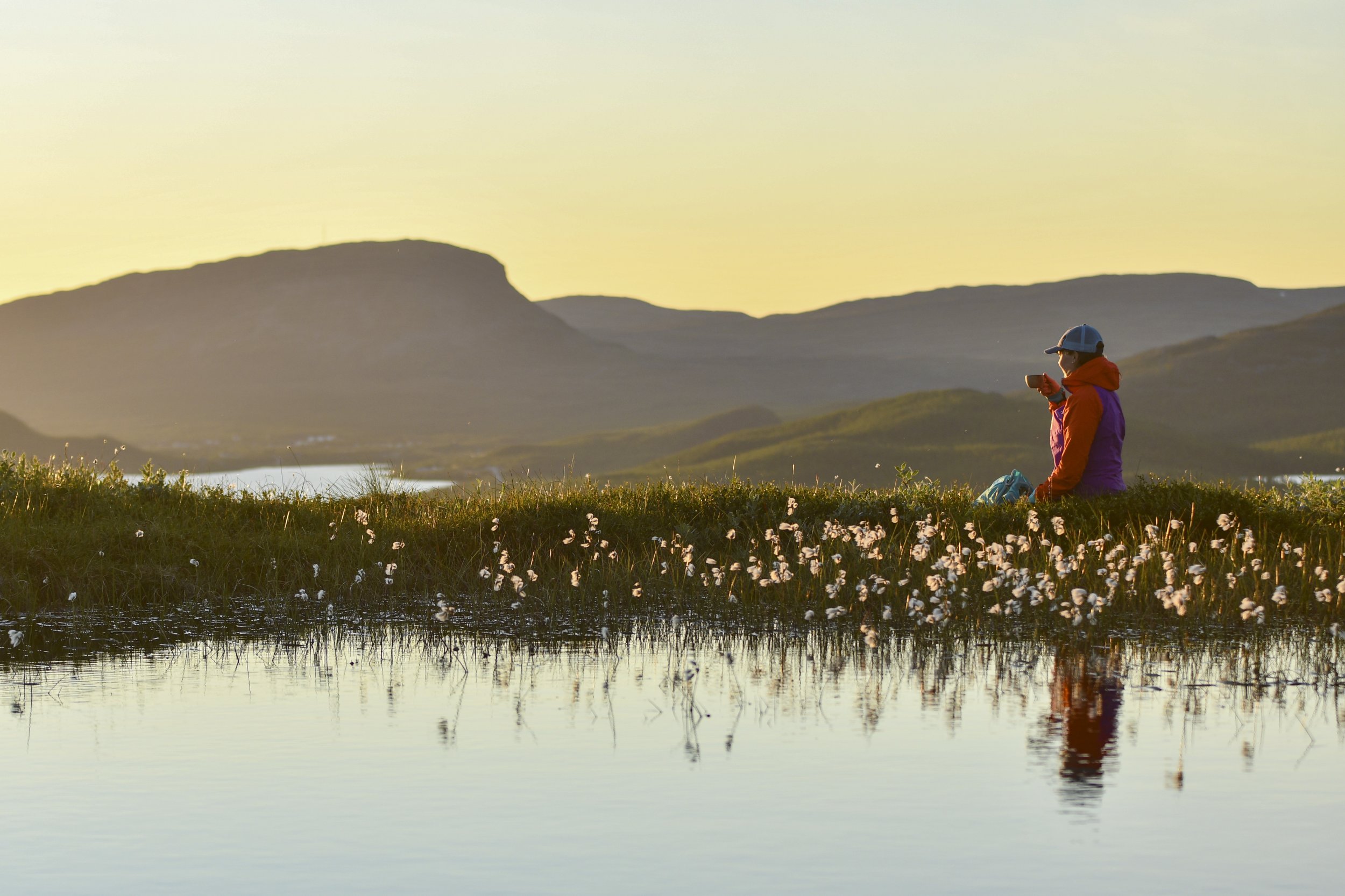 Kuvassa on kilpisjärven tunneituin nähtävyys, saanatunturi kesäyön valossa. kuvassa nainen istuu tunturilammen rannalla, juo kuksasta ja ihastelee upeaa maisemaa