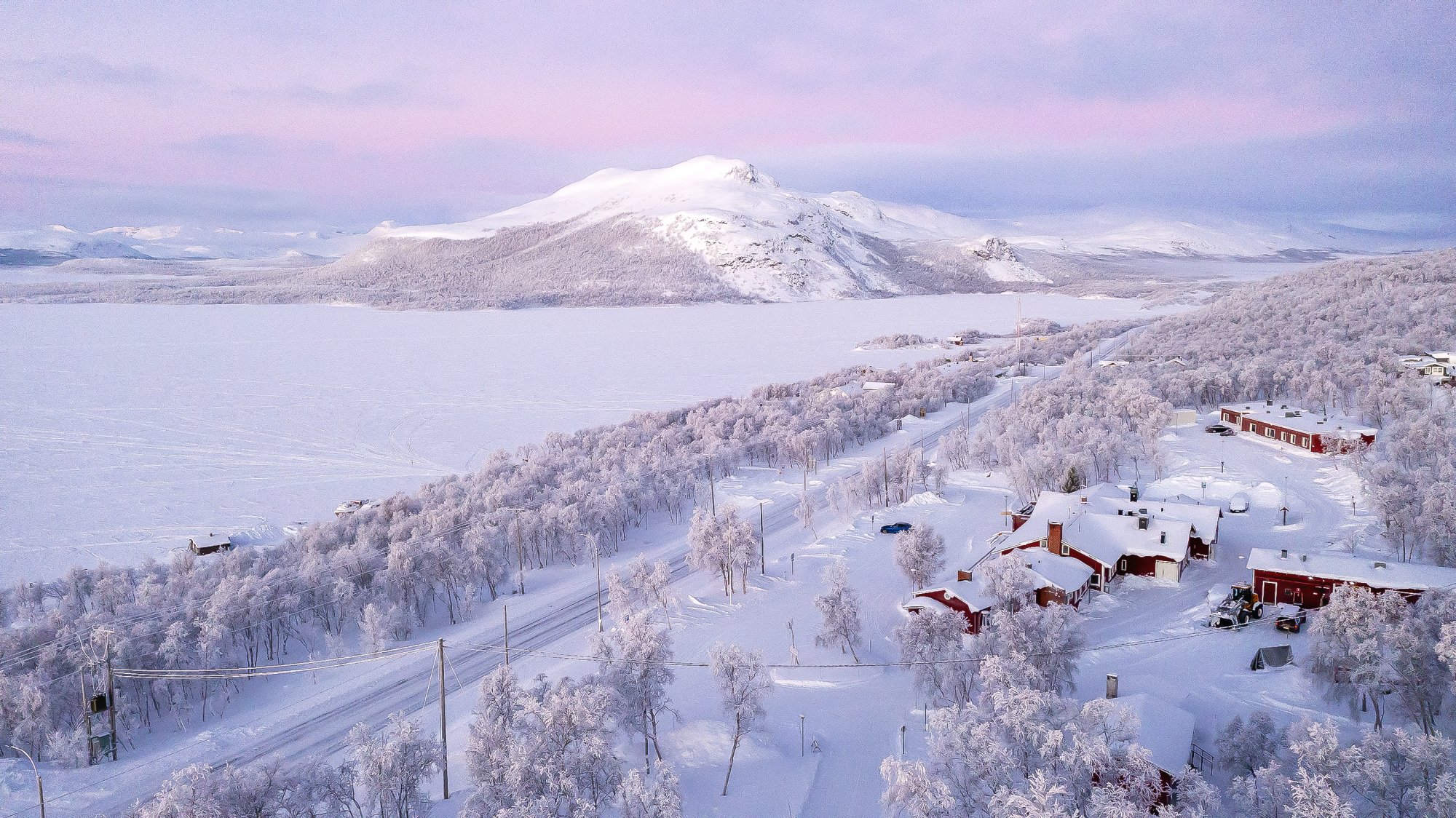 Kilpisjärven Retkeilykeskus kauniina, huurteisena pastellipäivänä. Rakennus sijaitsee lumisten puiden keskellä, taivas hehkuu pehmeän vaaleanpunaisena ja jäätynyt Kilpisjärvi avautuu sen edessä.