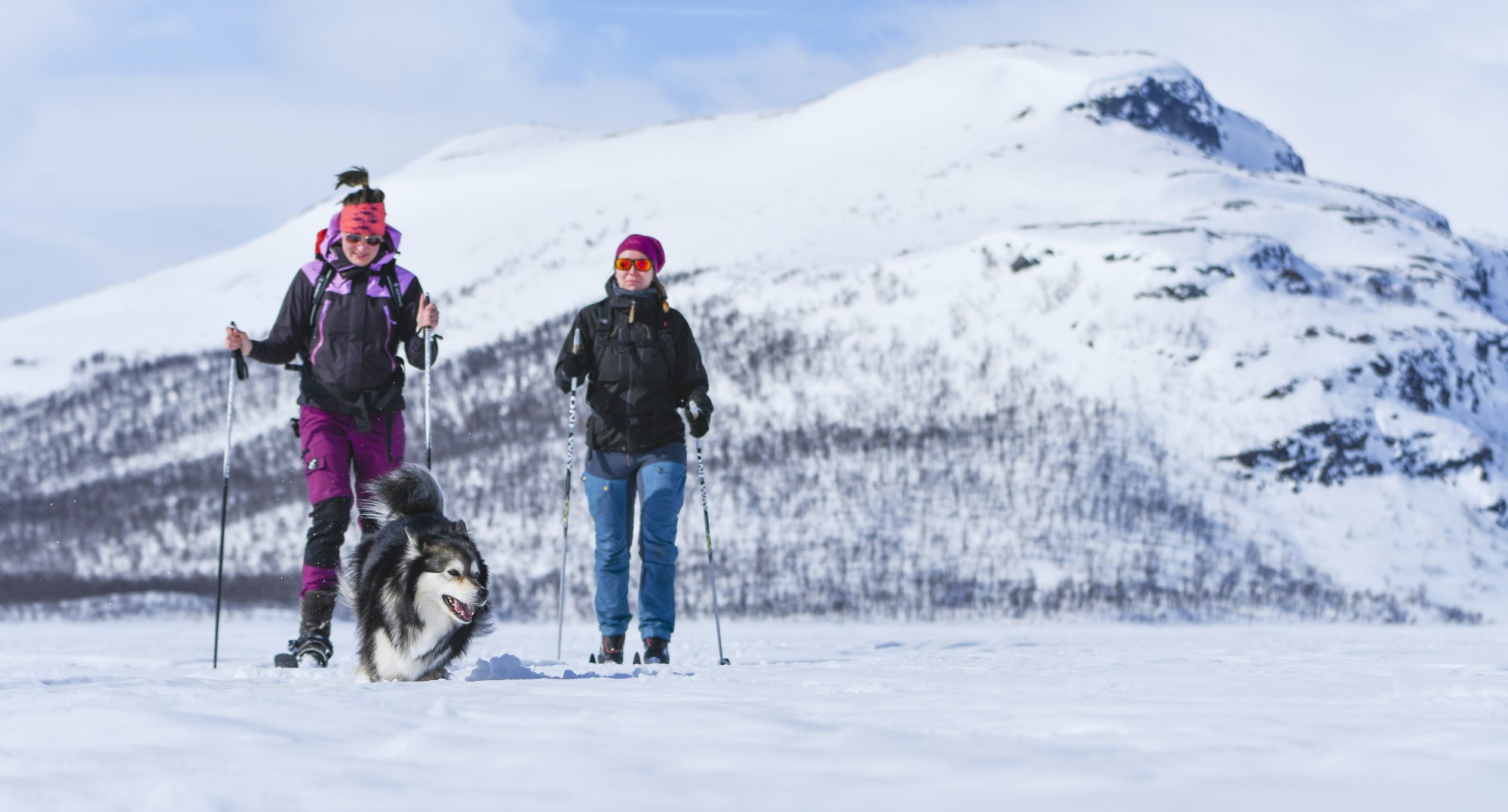 Kaksi naista hiihtää Kilpisjärven jäällä auringon paisteessa pikku-malla tunturi taustalla