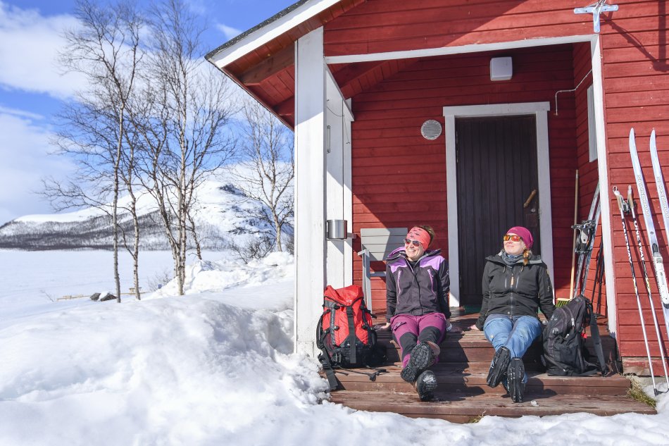 Punainen mökki Kilpisjärven rannalla keväisessä auringonpaisteessa. Kaksi naista ottaa portailla aurinkoa hiihtovaatteissa suksien nojatessa mökin seinään.