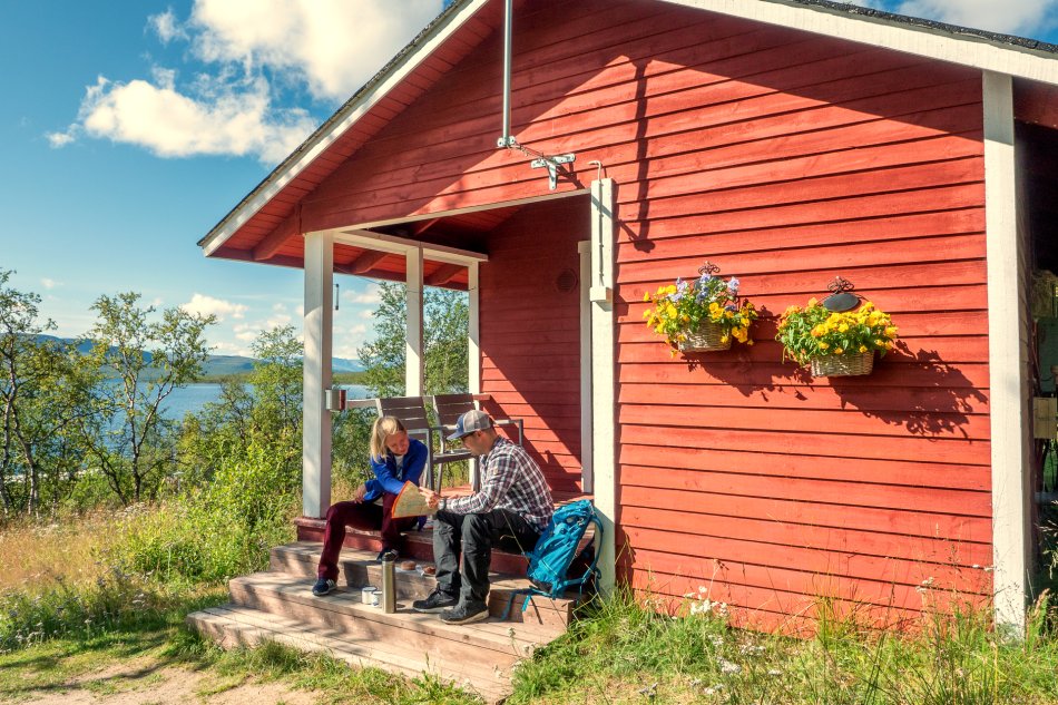 Punainen mökki Kilpisjärven rannalla keväisessä auringonpaisteessa. Kaksi naista ottaa portailla aurinkoa hiihtovaatteissa suksien nojatessa mökin seinään.