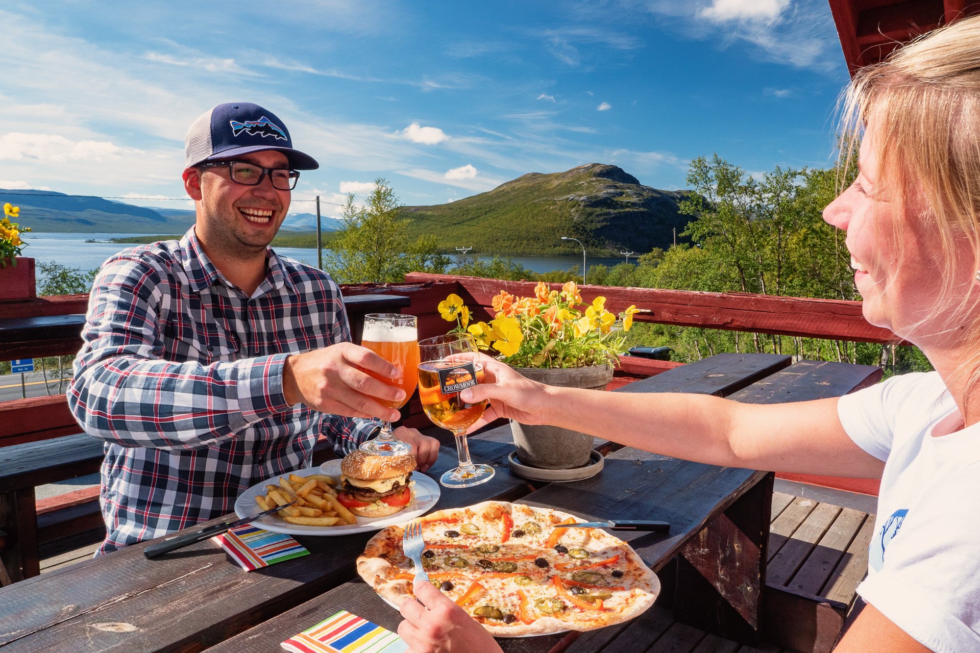 Kaksi ihmistä ruokailee ravintola Saanan terassilla Kilpisjärven Retkeilykeskuksella aurinkoisena kesäpäivänä. Näkymät ovat Kilpisjärvelle ja pikku-Mallalle. Pöydällä on pizza sekä hampurilainen ja kylmät juotavat