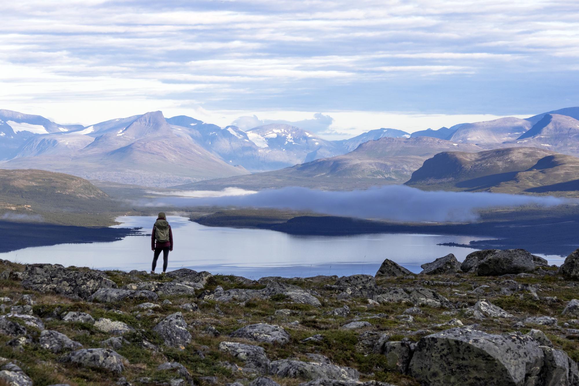 nainen seisoo selin kameraan Kilpisjärven tunturimaisemassa. Barras-tunturi näkyy kuvassa, samoin Mallan luonnonpuisto ja Kilpisjärvi. Aurinko nousee ja sumupilvi leijailee järven yllä. 