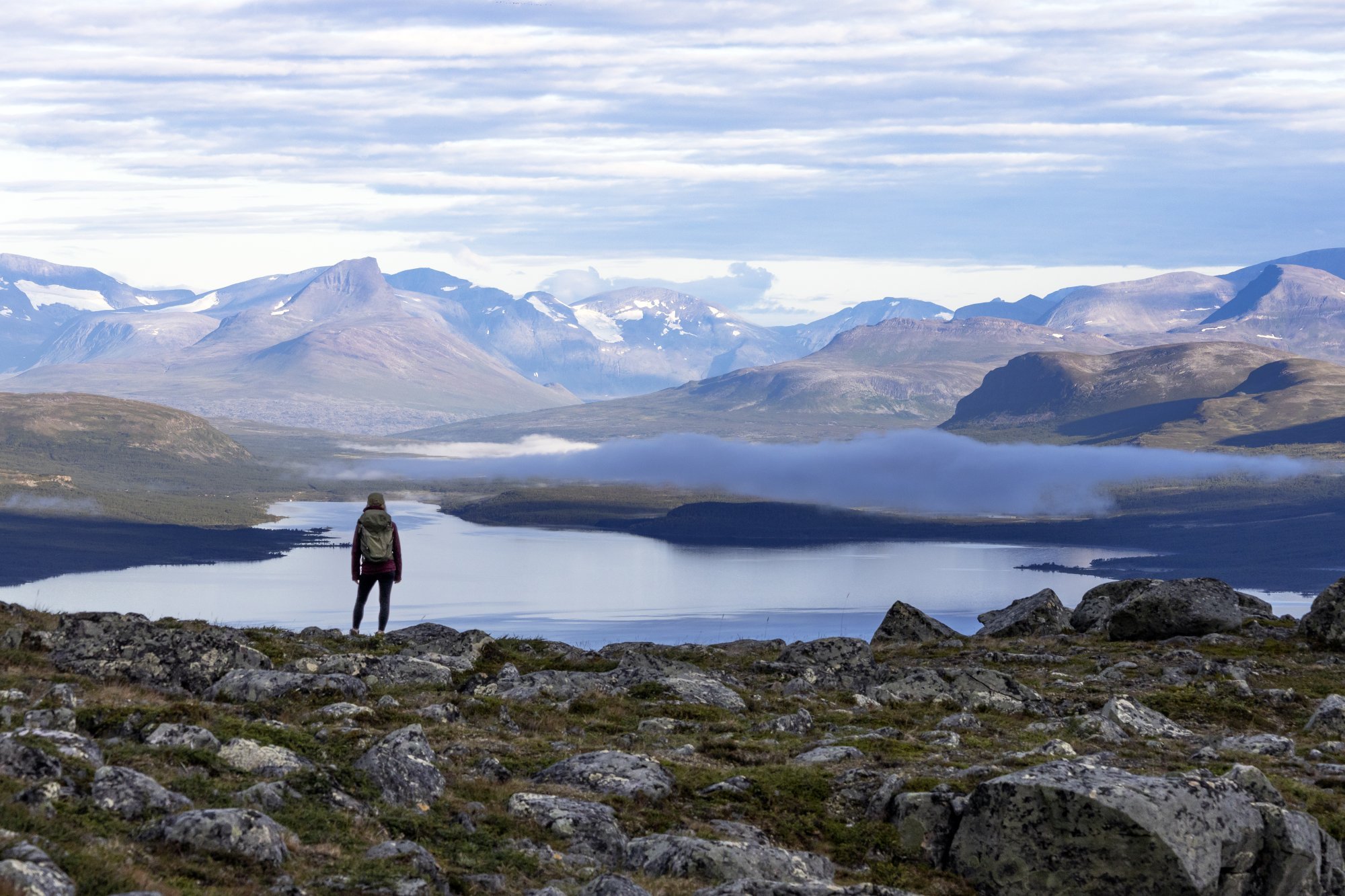 nainen seisoo selin kameraan Kilpisjärven tunturimaisemassa. Barras-tunturi näkyy kuvassa, samoin Mallan luonnonpuisto ja Kilpisjärvi. Aurinko nousee ja sumupilvi leijailee järven yllä. 